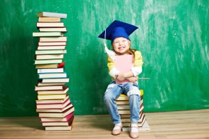 Joyful little girl wearing mortar board hat sitting on stack of books